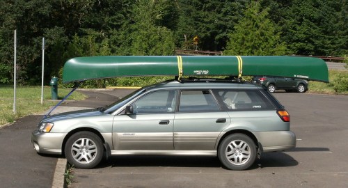 A silver station wagon parked with a green canoe secured on its roof, surrounded by trees in a parking area.