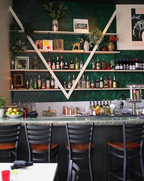 A stylish bar with green walls, shelves of liquor, and a sleek countertop, featuring empty chairs and decorative plants.