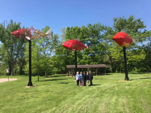 A group of people stands in front of large red fish sculptures on tall poles in a green park.