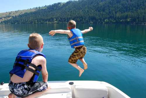 Two boys in life jackets, one jumping off a boat into a calm lake surrounded by trees and mountains.