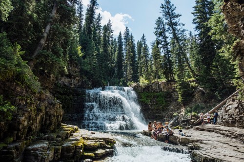 A serene waterfall surrounded by tall trees, with people sitting on rocks enjoying the natural beauty.