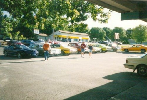 A group of classic cars parked in a lot, with people walking nearby and trees in the background.