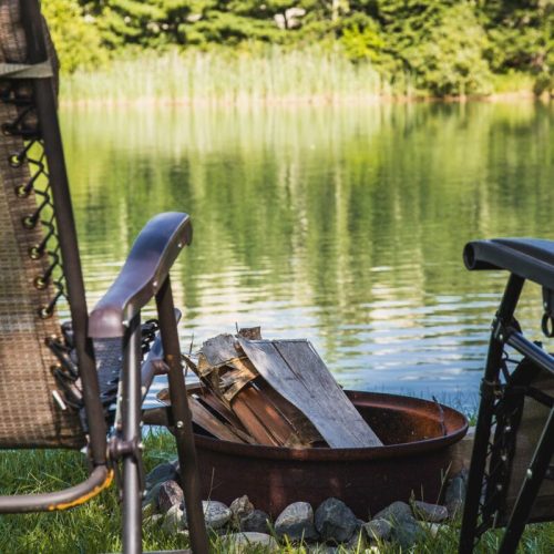 A serene lakeside scene featuring two chairs beside a fire pit with logs, surrounded by lush greenery and calm water.