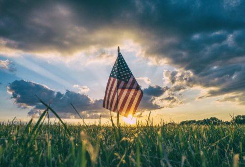 A small American flag stands in a grassy field, silhouetted against a sunset with dramatic clouds.