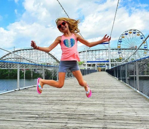 A girl in a pink tank top and sunglasses jumps joyfully on a wooden bridge with a carnival in the background.
