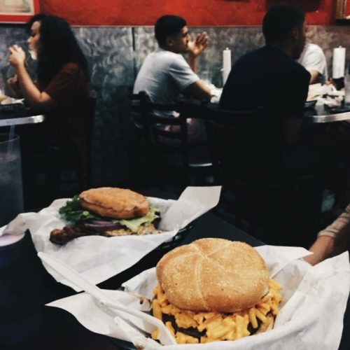 A close-up of a burger with mac and cheese on a table, with diners in the background enjoying their meals.