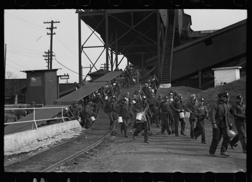 Black and white photo of workers in uniforms walking towards a coal mine, carrying buckets and tools.