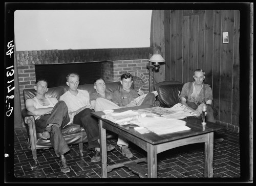 Five men sitting on a couch in a rustic room, reading and relaxing near a coffee table with newspapers.