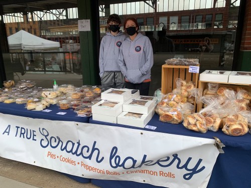 Two masked individuals stand behind a bakery table filled with various baked goods, including pies and cookies.