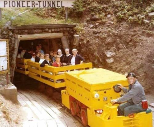 A yellow mining train with passengers enters the Pioneer Tunnel, surrounded by rocky terrain and greenery.