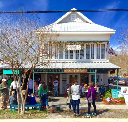A charming beach house restaurant with outdoor seating, people mingling, and a clear blue sky above.