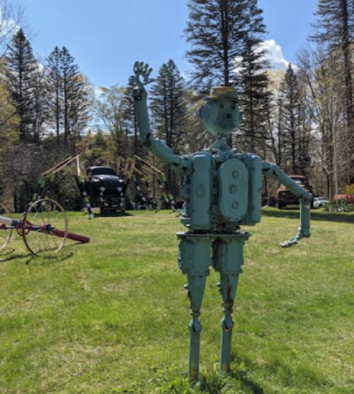 A whimsical robot sculpture made from metal parts stands in a grassy area, waving amidst trees and vintage machinery.