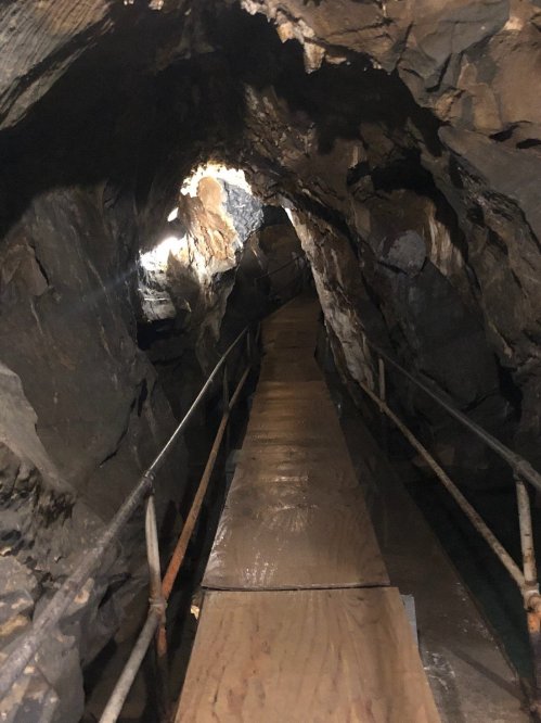 A narrow wooden walkway leads through a dark cave, flanked by rough stone walls and a faint light at the end.