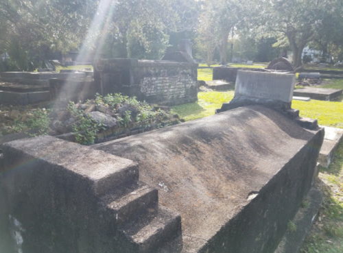 A weathered grave marker in a cemetery, surrounded by greenery and other gravestones under dappled sunlight.