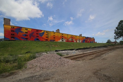 Colorful mural on a large building, with a grassy area and gravel path in the foreground, under a blue sky.