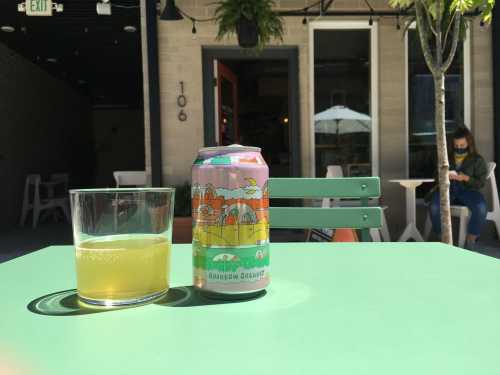 A colorful can of beer next to a glass on a green table, with a café setting in the background.