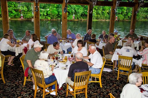 A lively restaurant scene with diners enjoying meals by a river, surrounded by greenery and wooden decor.