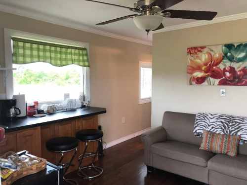 Cozy kitchen and living area with a sofa, bar stools, and floral artwork, featuring natural light from windows.