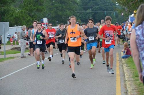A group of runners in athletic gear races down a road during a running event, with spectators in the background.