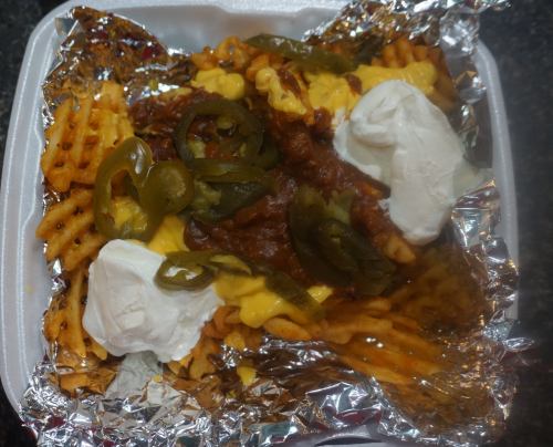 Waffle fries topped with cheese, chili, jalapeños, and dollops of sour cream, served in a foil container.