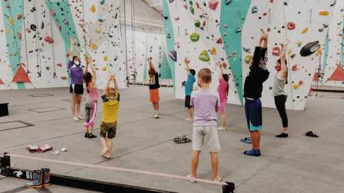 A group of children stretching in a climbing gym, surrounded by colorful climbing walls and equipment.