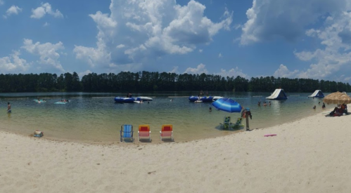 A sunny beach scene with people swimming, inflatable structures, and colorful chairs on the sand by a calm lake.