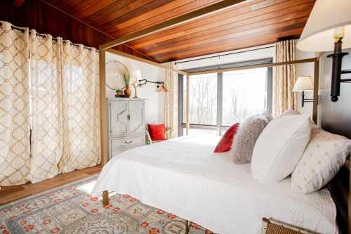 Cozy bedroom with a four-poster bed, large windows, patterned curtains, and warm wooden accents.