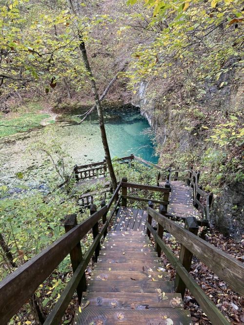 A wooden staircase descends through autumn foliage to a serene, turquoise pond surrounded by rocky cliffs.
