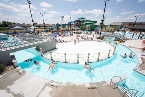 A sunny water park scene with a lazy river, water slides, and people relaxing by the pool.