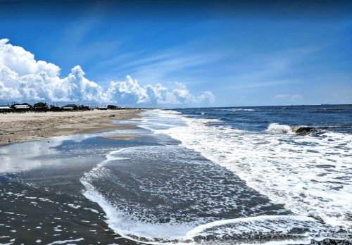 A serene beach scene with gentle waves, sandy shore, and a bright blue sky dotted with fluffy clouds.