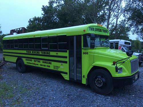A bright green bus labeled "Twin Rivers Tubing" parked on gravel, surrounded by trees.