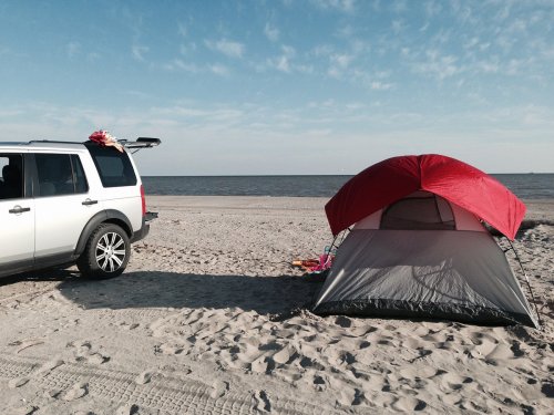 A silver SUV parked on a sandy beach next to a red and gray tent, with calm water and blue sky in the background.
