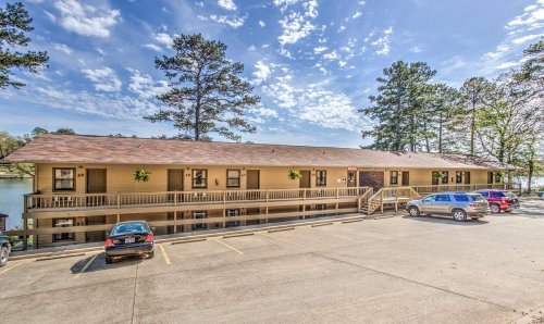 A lakeside motel with a wooden deck, surrounded by trees and parked cars under a blue sky with scattered clouds.