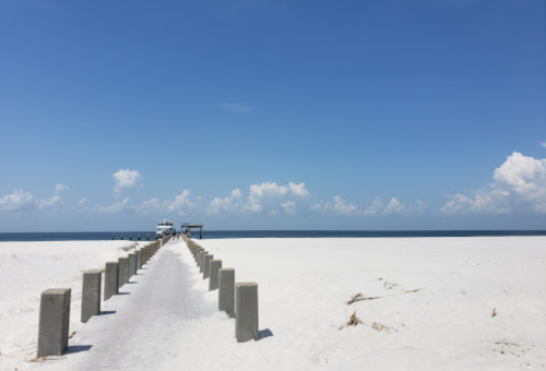 A long wooden pier extends over white sand towards the ocean under a clear blue sky with scattered clouds.
