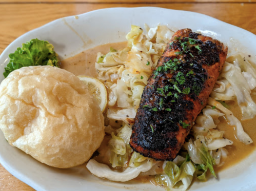 A plate of seasoned salmon on a bed of sautéed cabbage, served with a fluffy bread roll and a lemon wedge.