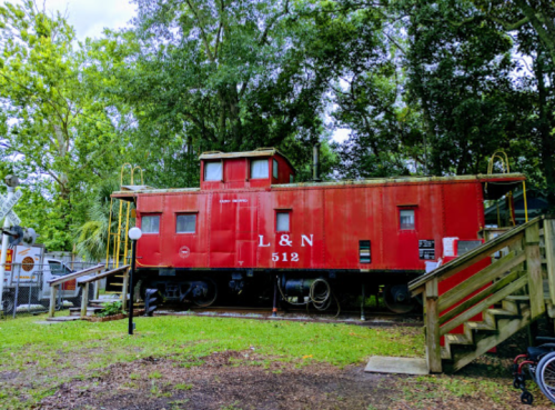 A red caboose labeled "L&N 512" sits among trees, with stairs leading up to it in a grassy area.