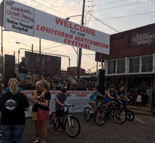 A lively street scene at the Louisiana Watermelon Festival, featuring a stage, vendors, and people on bicycles.