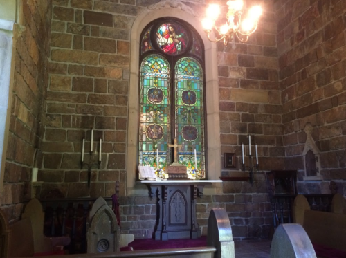 Interior of a stone church featuring a stained glass window, candles, and wooden pews.
