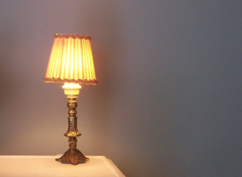 A vintage lamp with a pleated shade sits on a table against a blue wall. The light is softly glowing.