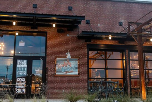 Exterior of a brick building with large windows, featuring a sign that says "Coming Soon" and outdoor seating.