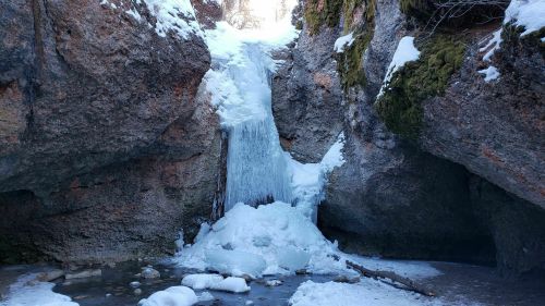 A frozen waterfall surrounded by rocky cliffs and snow, with ice formations at the base and a small pool of water.