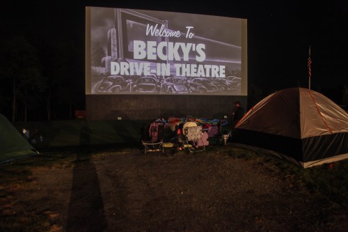 A drive-in theater screen displays "Welcome to Becky's Drive-In Theatre" with tents and chairs set up in the foreground.