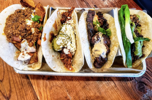 A platter of four tacos with various fillings, including meat, cheese, and toppings, served on a wooden table.