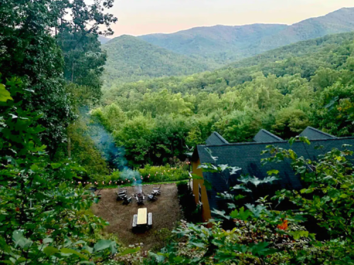 A scenic view of mountains and lush greenery, with a fire pit area and a house nestled among the trees.