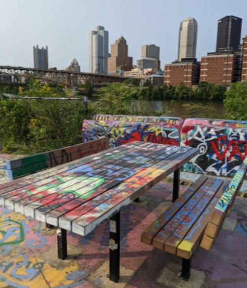A colorful, graffiti-covered picnic table overlooks a city skyline by a river.