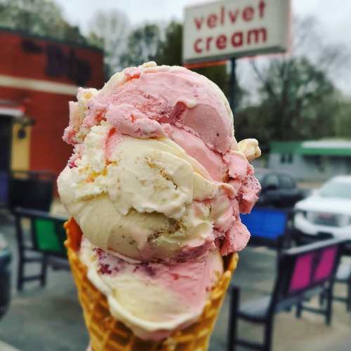 A close-up of a colorful ice cream cone with pink, white, and yellow scoops, with a shop sign in the background.