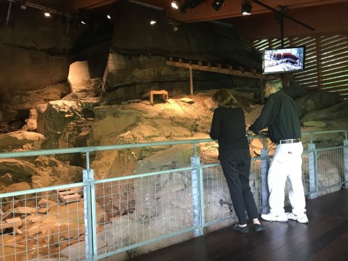 Two visitors observe an archaeological site from a viewing platform, with rocks and a display screen in the background.