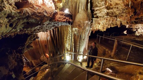 A person stands on a staircase in a cave, surrounded by illuminated stalactites and stalagmites.
