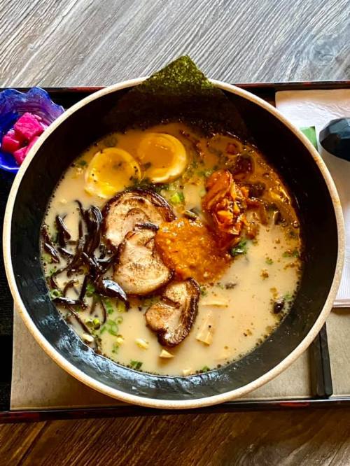 A bowl of ramen with broth, sliced pork, soft-boiled eggs, mushrooms, and garnishes, served on a wooden table.