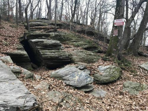 Rocky terrain with large boulders, scattered leaves, and trees in a wooded area. A warning sign is visible nearby.
